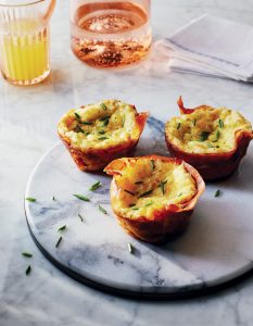 Three egg muffins garnished with chives sit on a round marble platter. A glass of orange juice, a folded napkin, and a glass with a pink beverage are in the background on a marble surface.