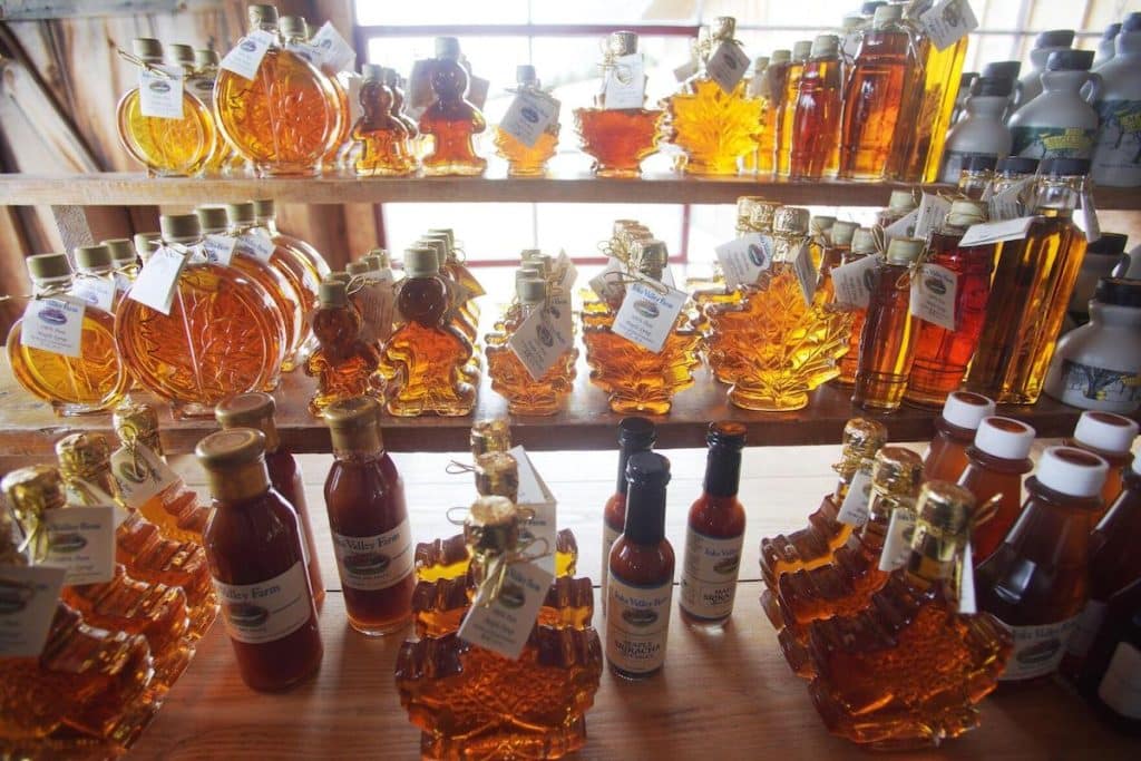 Various shapes and sizes of maple syrup bottles are displayed on wooden shelves in front of a window. The bottles range from small to large, featuring different designs such as leaves and animals.