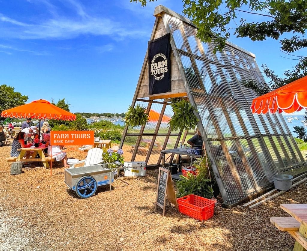 Outdoor farm tour base camp with a triangular greenhouse structure, various plants, orange umbrellas over picnic tables, and a farm tours sign by the entrance.