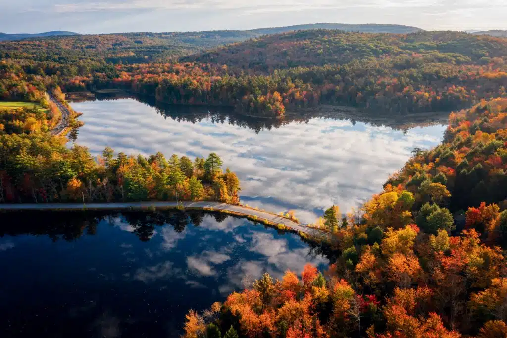 Aerial view of a forested landscape in autumn with a heart-shaped lake and reflected clouds, surrounded by colorful trees and hills.