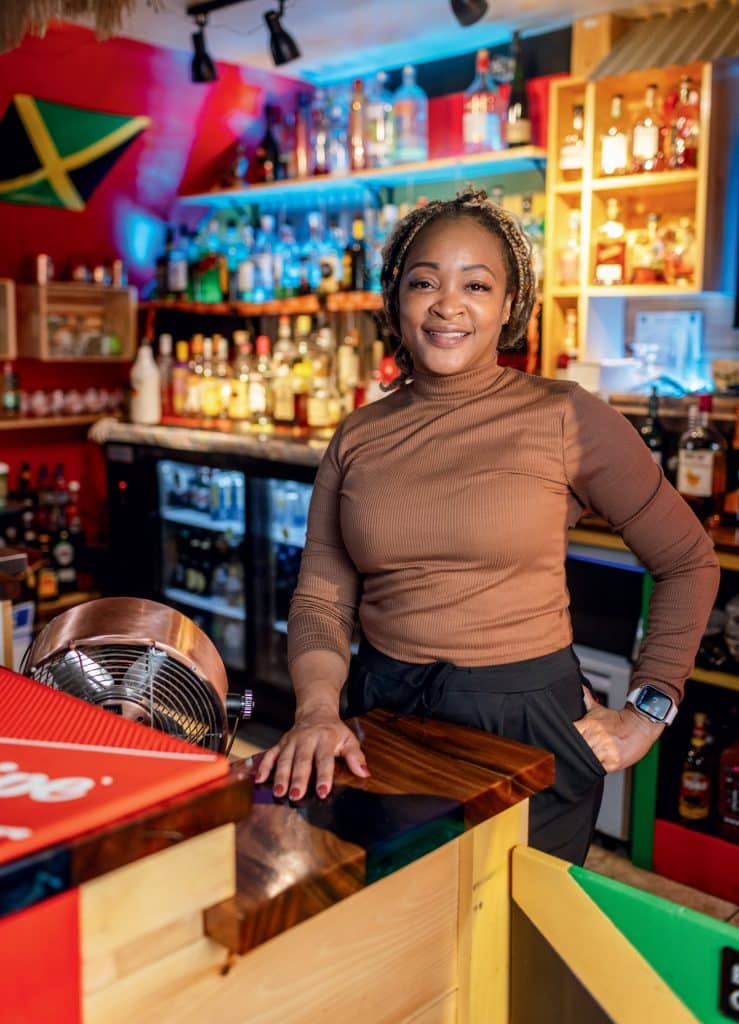 A woman stands behind the counter of a colorful bar with shelves of liquor bottles in the background. A Jamaican flag hangs on the wall.