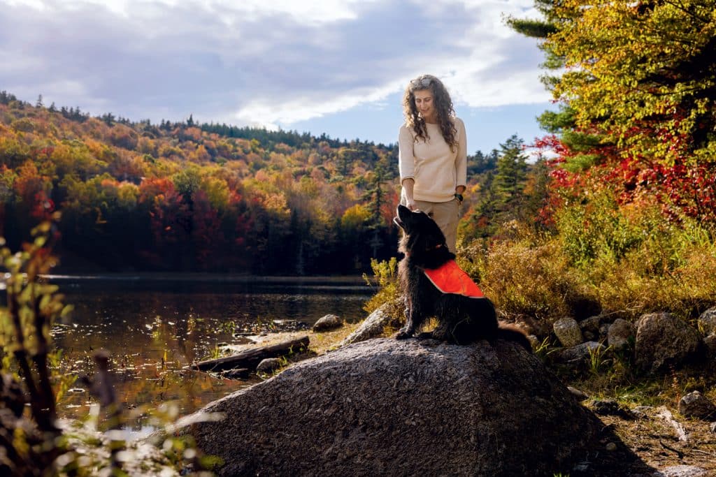 A person with curly hair stands beside a dog wearing an orange vest, both on a large rock by a lake, with a forest displaying vibrant fall colors in the background.