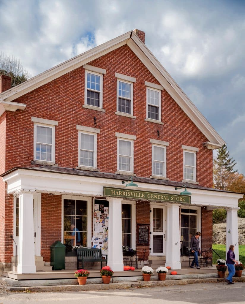 Red brick building with white trim labeled "Harrisville General Store". Potted plants and pumpkins on the porch. Two people are sitting with one standing in front. Cloudy sky in the background.