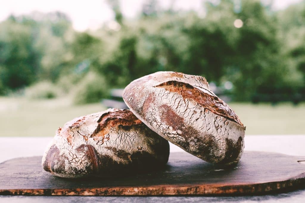 Two round, rustic loaves of bread with a crusty exterior sit on a wooden board. A blurred outdoor background with greenery is visible behind them.