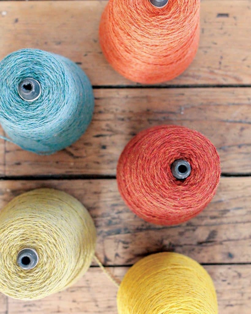 Five spools of yarn in blue, orange, red, yellow, and light yellow are arranged on a wooden surface.