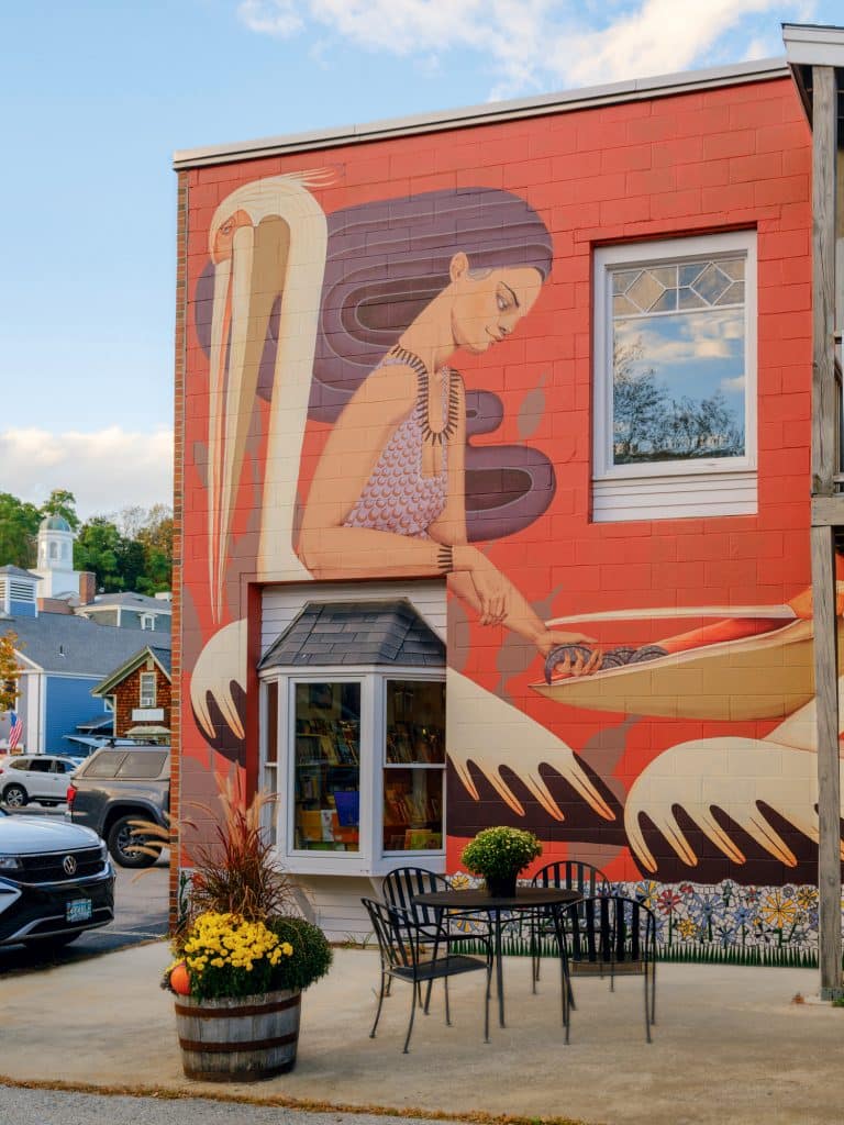 A large mural of a woman cooking covers the side of a brick building. In front, there is an outdoor seating area with a table, chairs, and a planter with flowers.