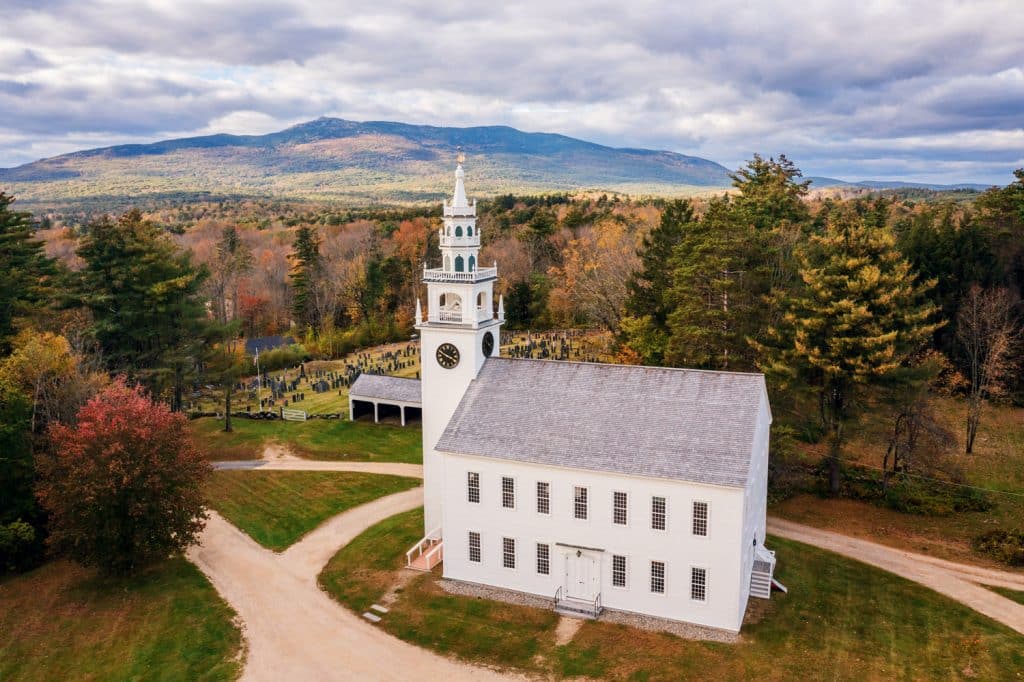 Aerial view of a white church with a clock tower, situated in a rural area with surrounding trees and a cemetery, against a backdrop of mountains under a partly cloudy sky.