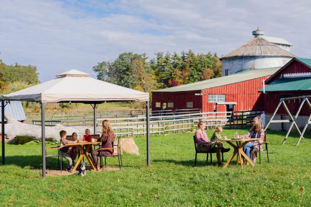 People sitting at tables under a canopy and outside it on a sunny day at a farm with red buildings and a silo in the background.