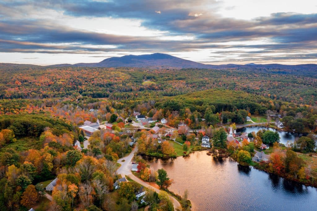 Aerial view of a rural landscape in autumn, featuring a town, colorful trees, a lake, and mountains in the background under a cloudy sky.