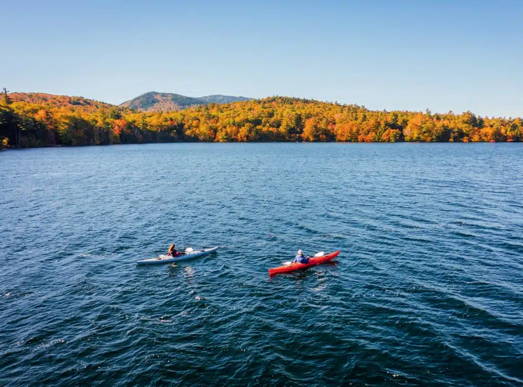 Two people kayaking on a serene lake with forested hills displaying autumn colors in the background under a clear blue sky.