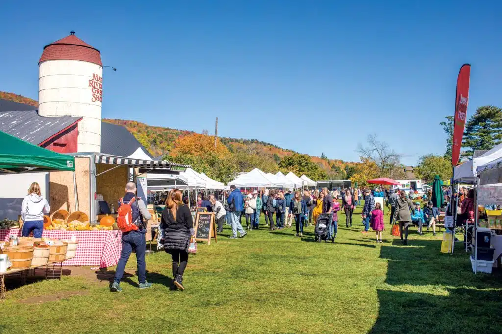 People browsing stalls at an outdoor farmers market on a sunny day near a barn with a silo, surrounded by trees with autumn foliage.