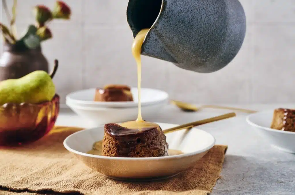 Person pours sauce over a small cake on a plate with a spoon beside it; pear and another plated cake in the background, for Pear Sticky Toffee Puddings