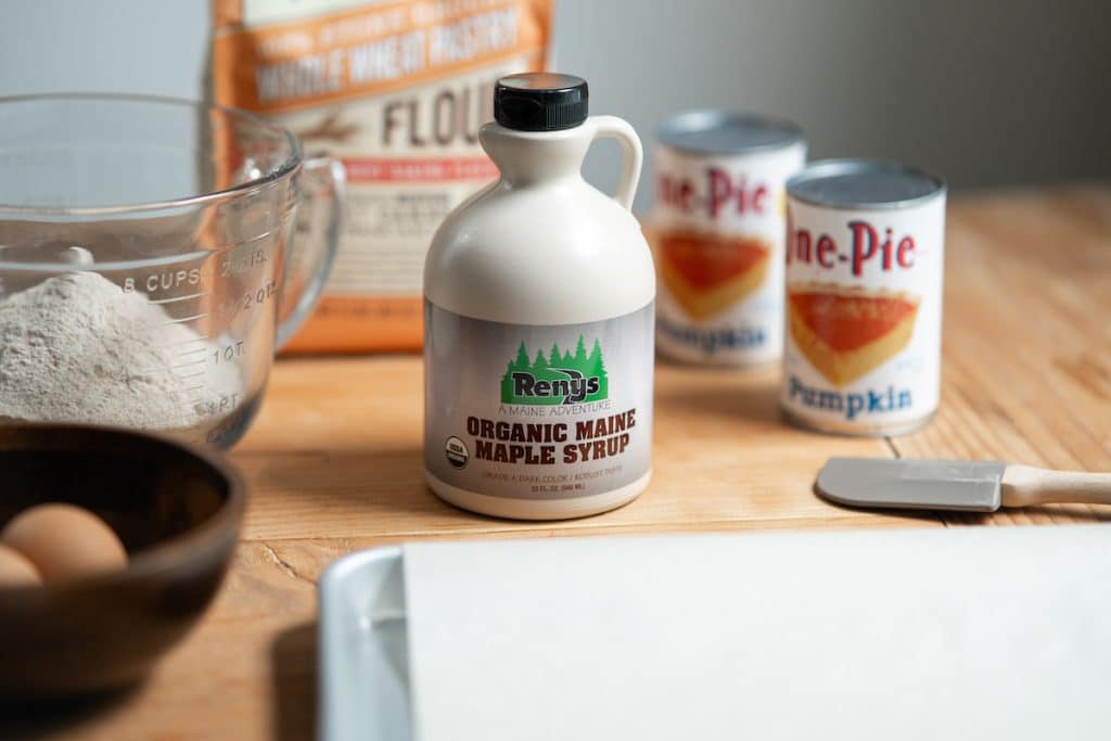 A bottle of organic Maine maple syrup on a wooden table surrounded by baking ingredients: flour, canned pumpkin, eggs, and a spatula next to a baking sheet.