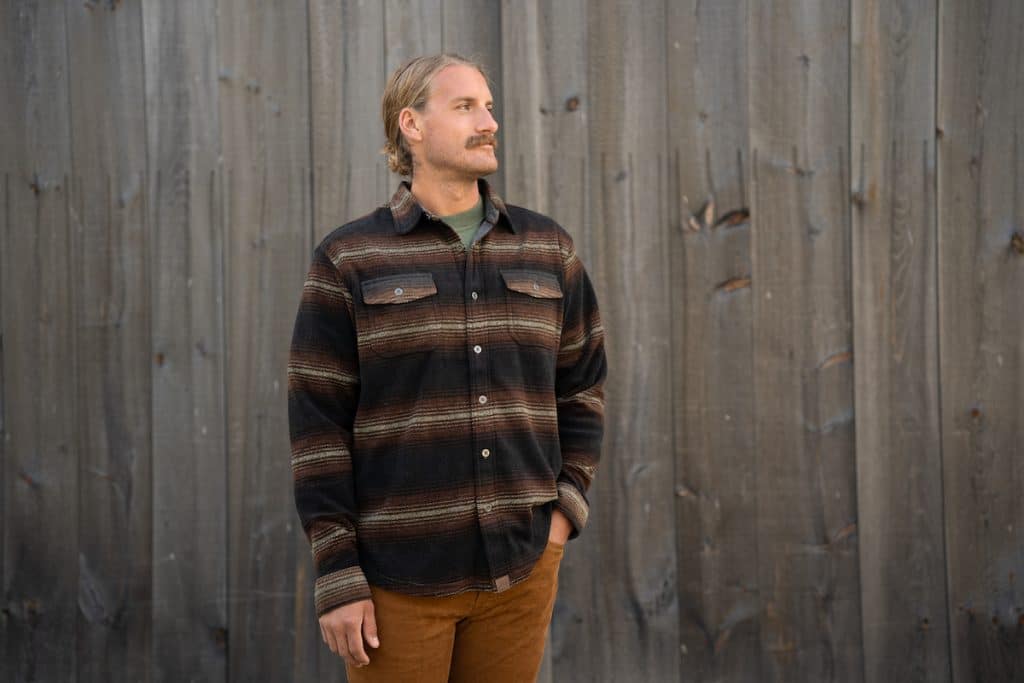 A man with long hair and a mustache, wearing a striped shirt and brown pants, stands in front of a wooden wall, looking to the side.