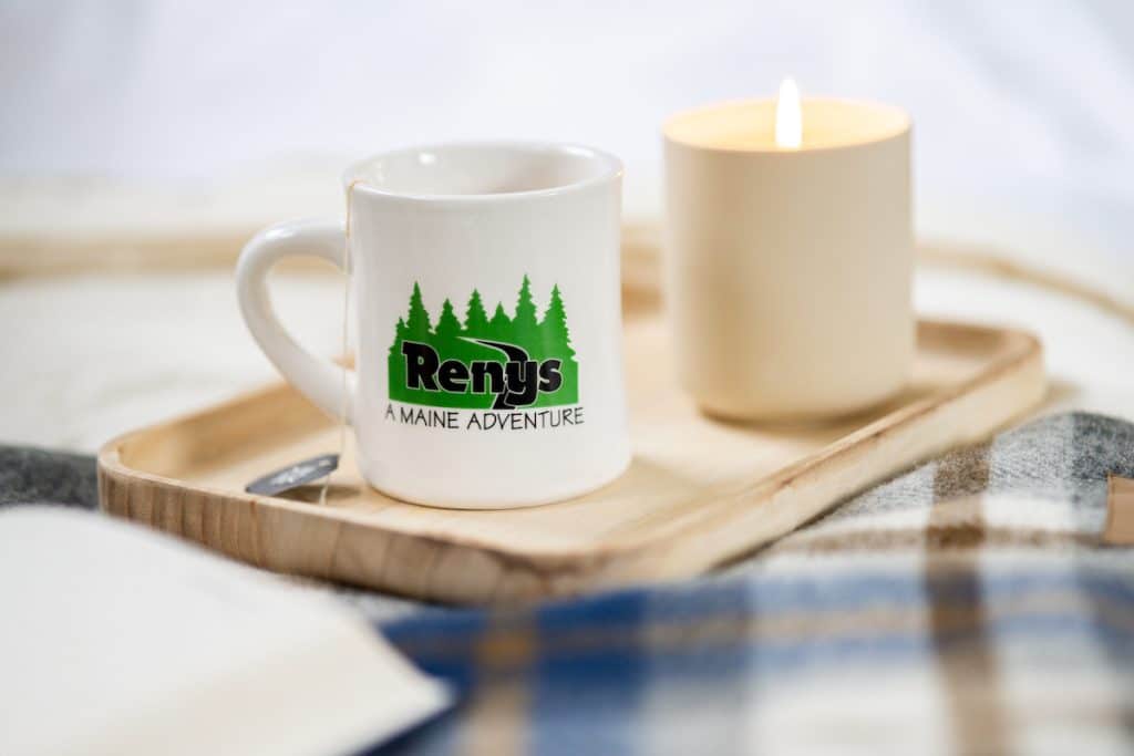 A white mug with the text "Renys: A Maine Adventure" and tree graphics sits on a wooden tray next to a lit candle. A cozy blanket is visible underneath.