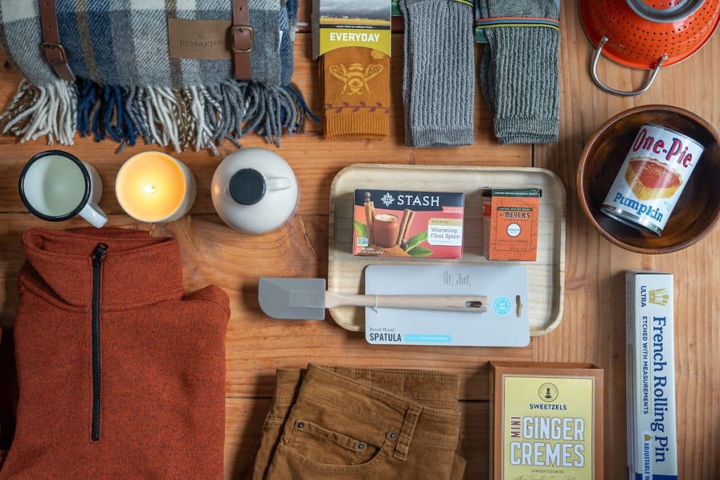 An assortment of items on a wooden surface including clothing, a blanket, tea, candles, a spatula, baking ingredients, socks, and a French Rolling Pin.