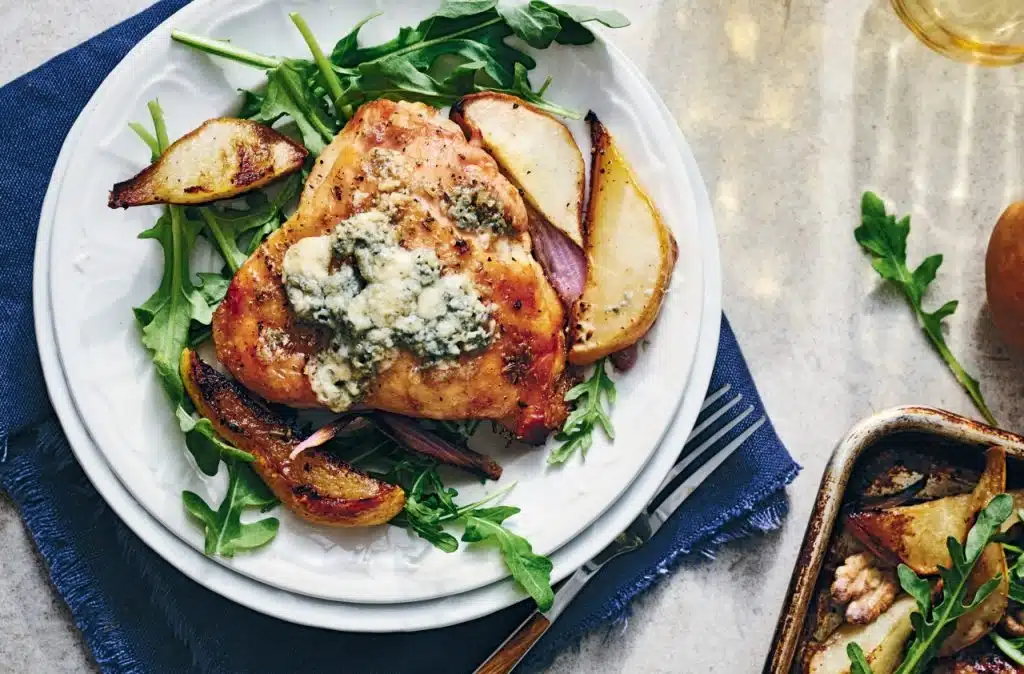 Sheet Pan Chicken with Pears, Onion, Walnuts, and Blue Cheese. Plate of roasted chicken topped with blue cheese crumbles, served on a bed of arugula with roasted pears and onions, next to a small tray of roasted vegetables.