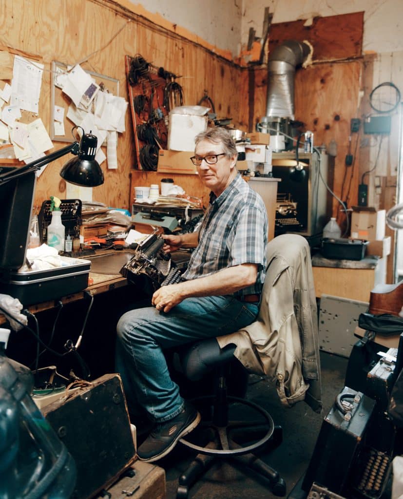 A person sits at a cluttered workbench in a workshop, surrounded by various tools, papers, and equipment.