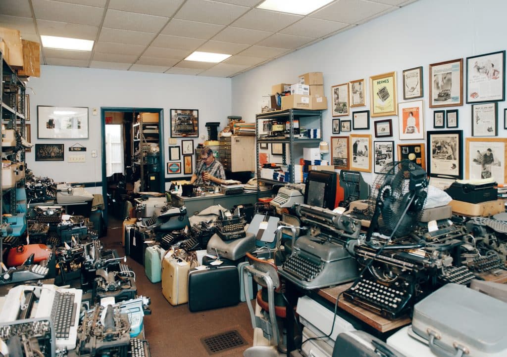 A cluttered room filled with numerous vintage typewriters, various office supplies, and framed pictures on the walls. A person sits at a desk, partially visible in the background.