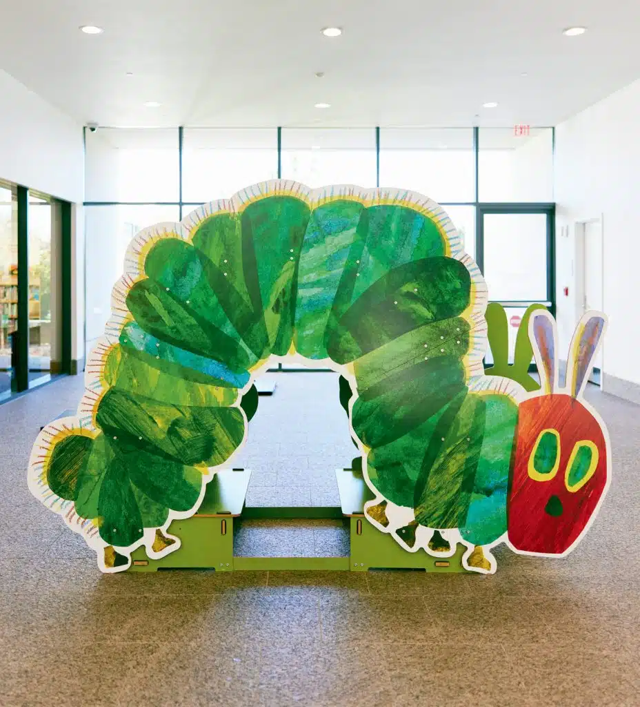 A large, vibrant cutout of a green caterpillar with a red head from the children’s book "The Very Hungry Caterpillar" is displayed in a brightly lit indoor area.