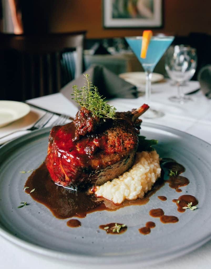 A gourmet dish of glazed meat on a bed of risotto, garnished with herbs, is served on a grey plate with a blue cocktail in the background.