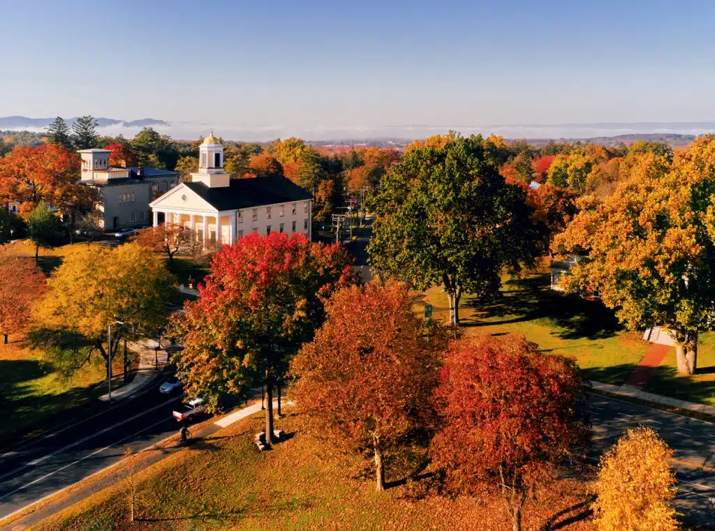 Aerial view of a building with a white cupola surrounded by autumn foliage, trees in shades of red, orange, and yellow, clear sky, and distant mountains.