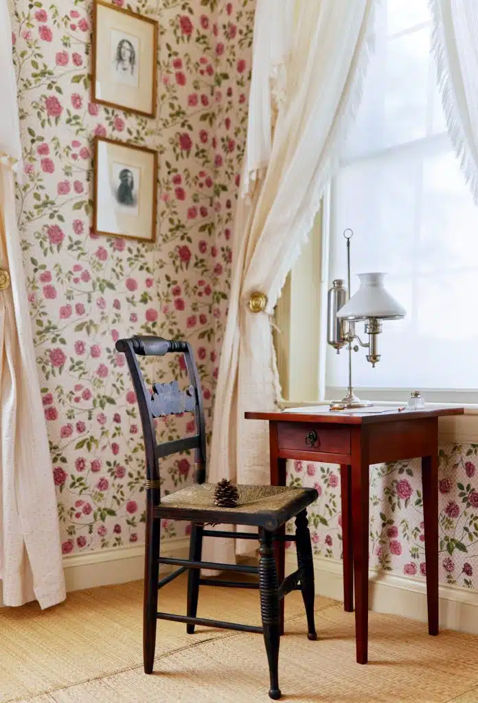 A room with floral wallpaper, a black wooden chair, a table with a lamp, and two framed pictures on the wall. White curtains drape over a sunlit window. A pine cone lies on the chair.