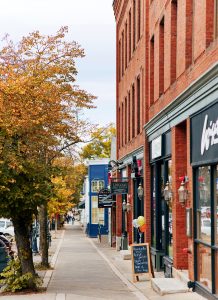 A narrow sidewalk lined with red brick buildings and storefronts on a clear autumn day. Trees with golden leaves are visible along the street, and a few pedestrians are seen in the distance.