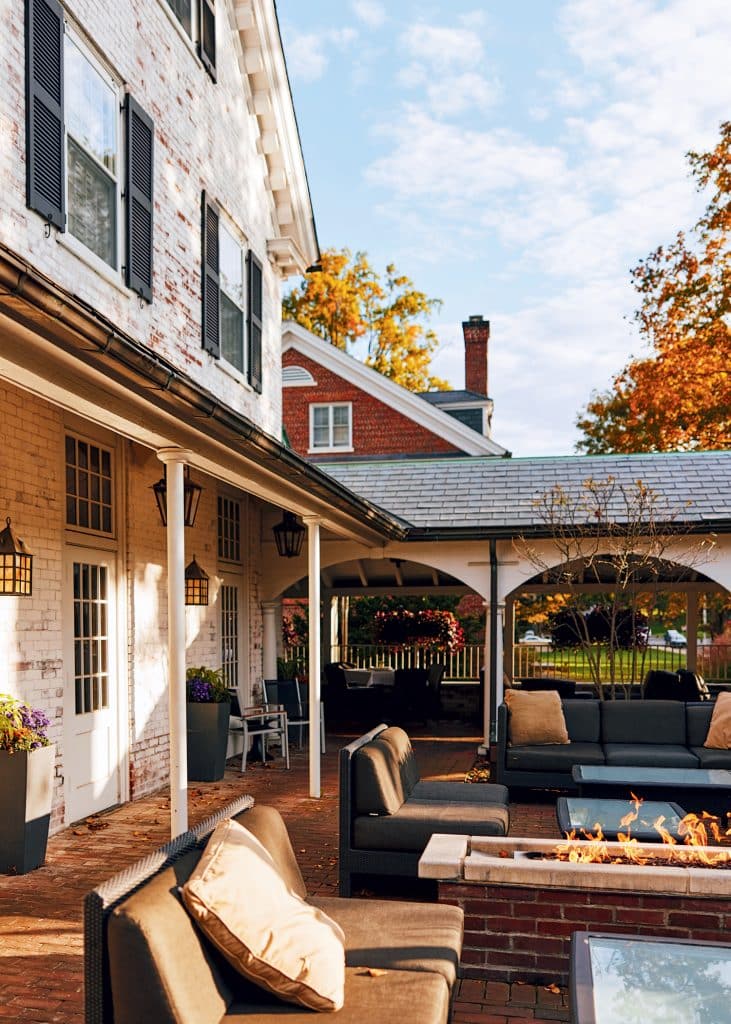 Outdoor patio with cushioned seating, a fire pit, and architectural overhang. The colonial-style building features white brick and dark shutters. Autumn trees are visible in the background.