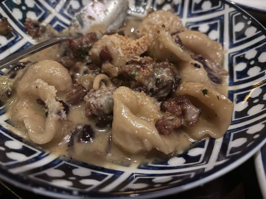 Close-up of a bowl containing a creamy pasta dish with mushrooms, ground meat, and breadcrumbs. The bowl has a blue and white geometric pattern. A spoon rests inside the bowl.