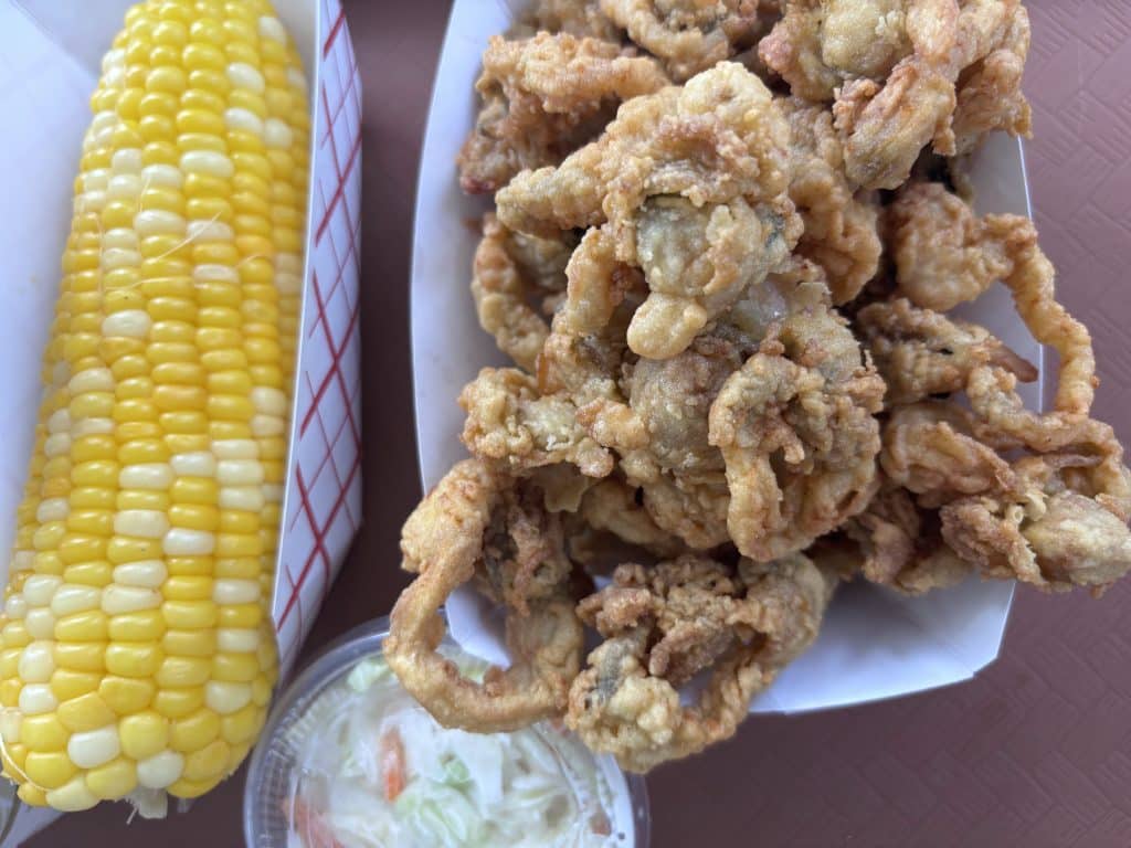 A serving of fried seafood in a paper tray, accompanied by a cob of corn and a portion of coleslaw in a small container.