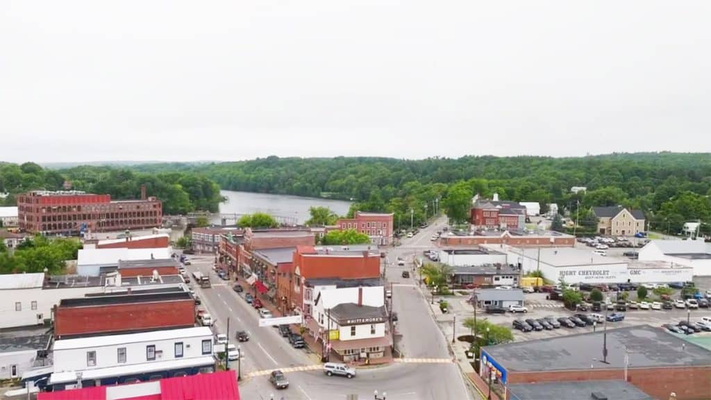 Aerial view of a small town with a river in the background. The image shows roads, parked cars, and various buildings, including brick structures and a white industrial building. Trees line the horizon.