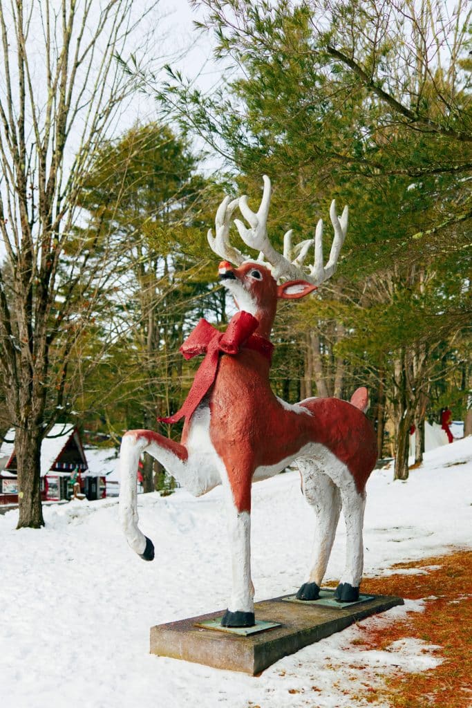 A large reindeer statue with a red bow is displayed outdoors on a snowy day, surrounded by trees.