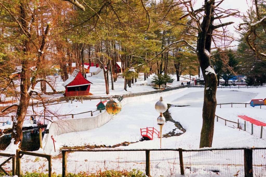 Snow-covered park with trees, a red building, and a small bridge. Fenced path and scattered playground equipment are visible.