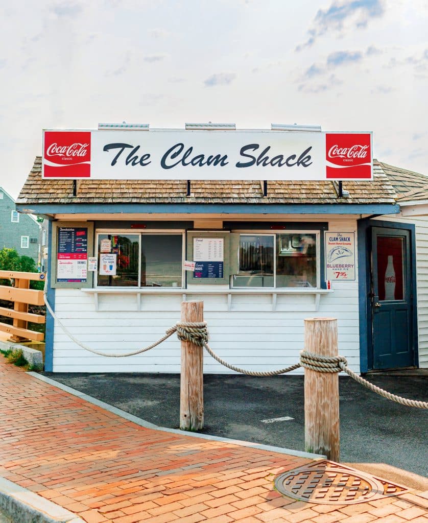 The photo shows a small, white seafood shack with a sign reading "The Clam Shack." It features Coca-Cola branding and a brick walkway with rope barriers in front.