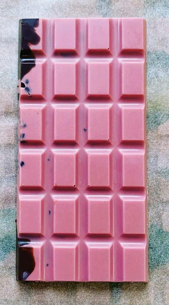 A pink chocolate bar with a grid pattern, featuring some dark spots in the top left corner, placed on a textured surface.