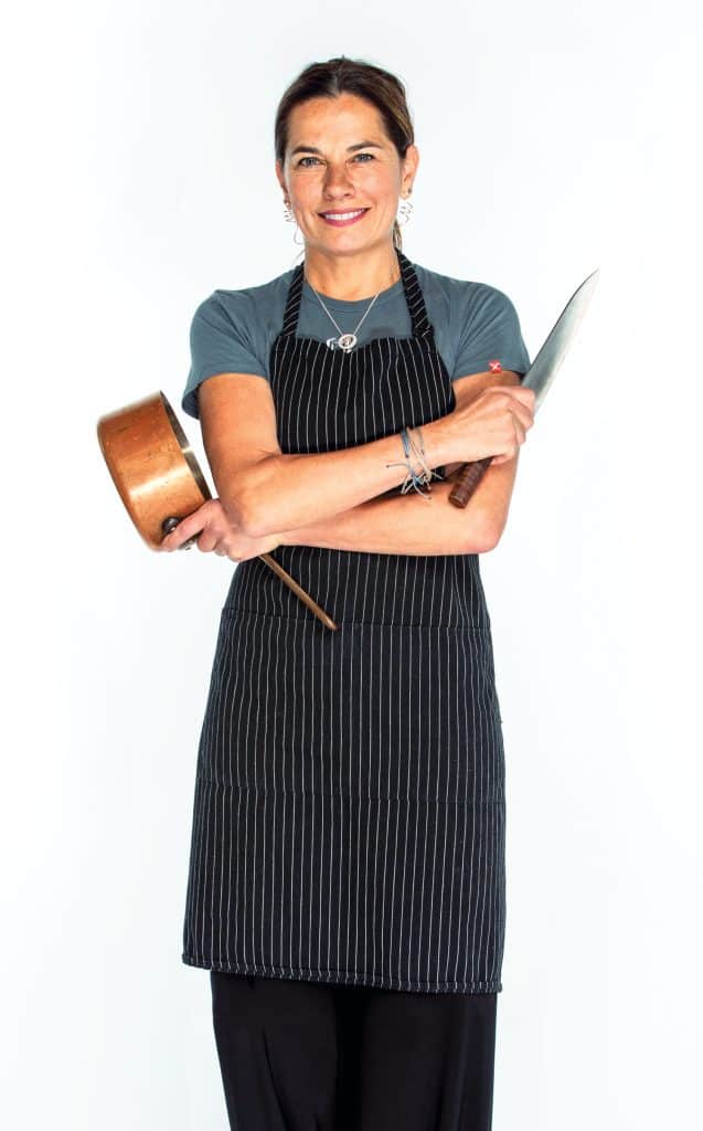 A person wearing a striped apron holding a saucepan and a knife, standing against a plain background.