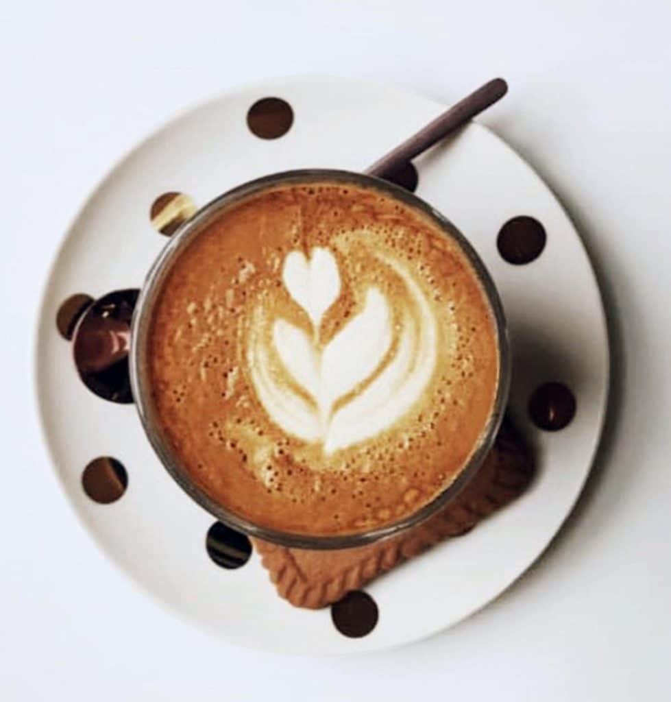 A cup of coffee with latte art and a biscuit on a white polka dot saucer.