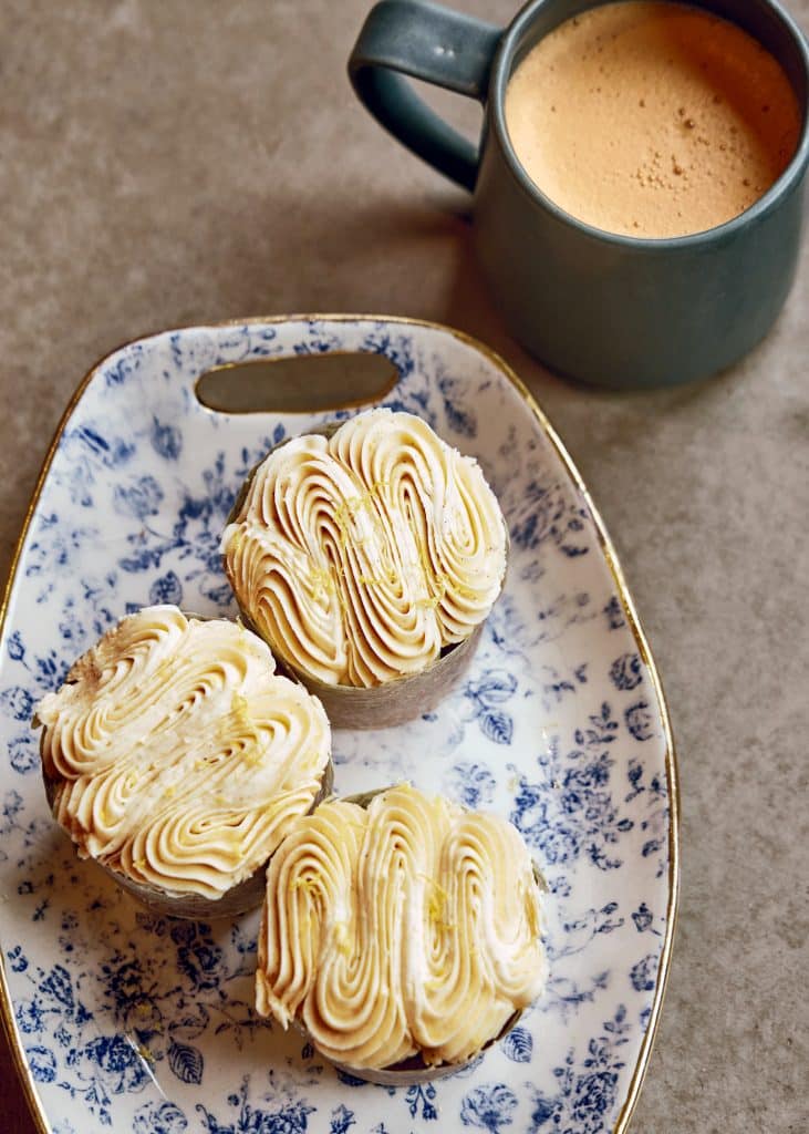 Three cupcakes with swirled frosting on a floral plate next to a cup of coffee.