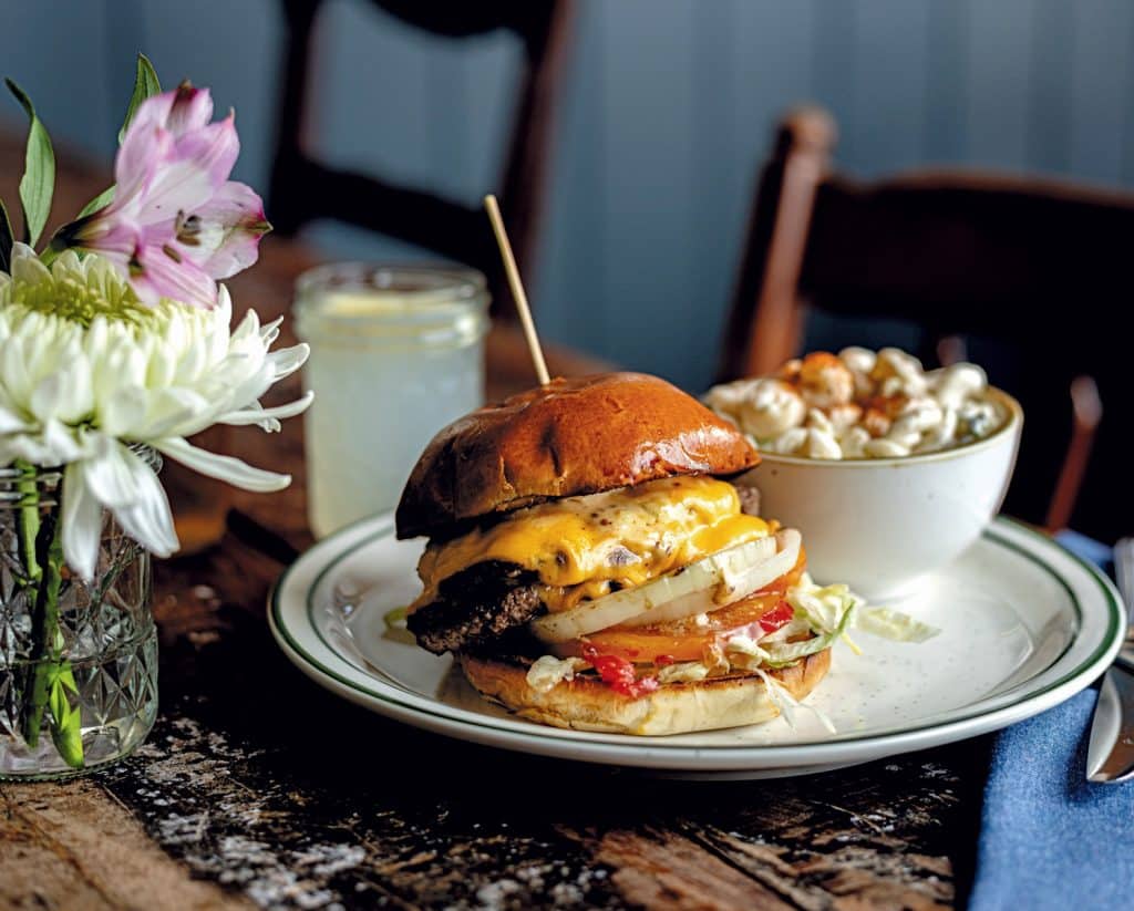 A cheeseburger with lettuce and tomato on a plate, accompanied by a bowl of salad. A drink and a vase with flowers are in the background.