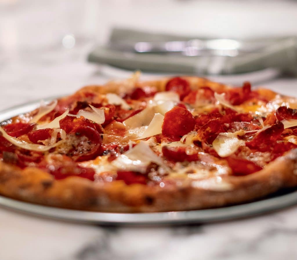 A pepperoni pizza topped with cheese and herbs on a metal plate, placed on a marble table. In the background, there are blurred napkins and silverware.