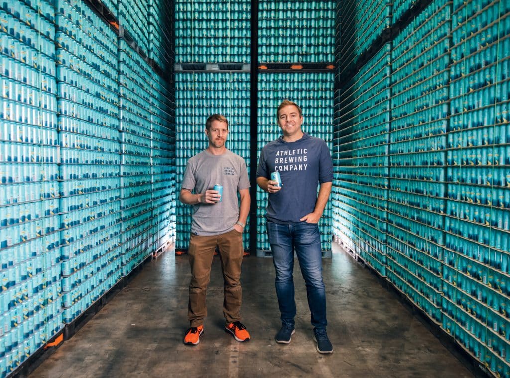 Two people stand in a warehouse stocked with tall stacks of blue beverage cans, each holding a beverage can.