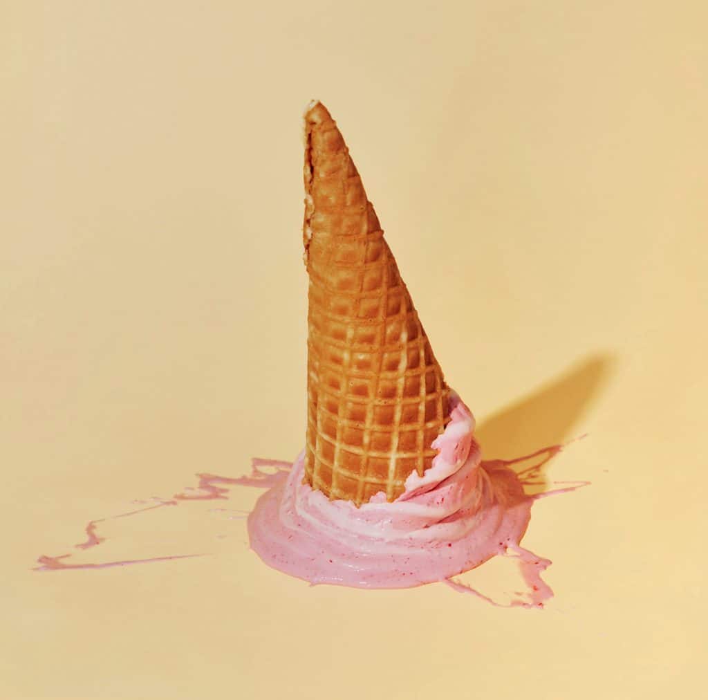 A waffle cone placed upside-down in a pool of melted pink ice cream on a yellow surface.