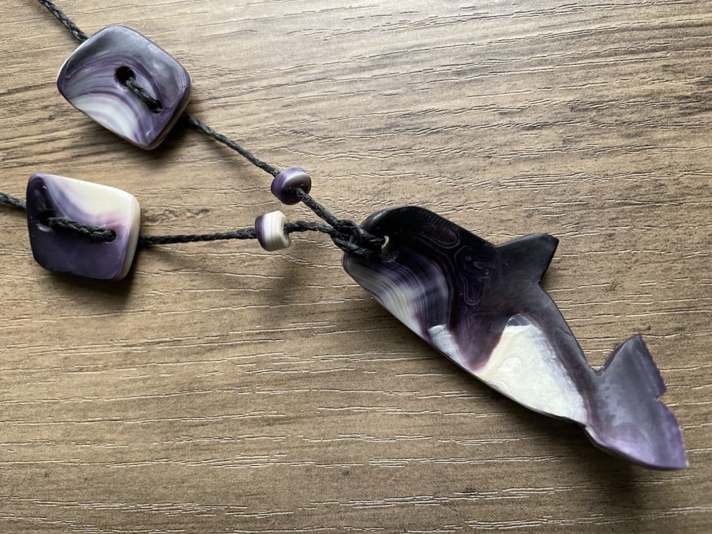 A whale-shaped pendant made of multicolored stone, attached to a black string necklace with two square beads and small round accents, on a wooden surface.