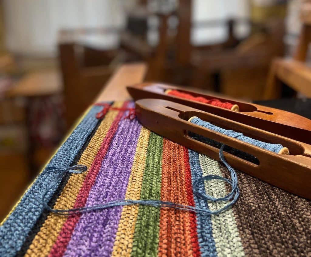 Colorful woven fabric on a loom with two wooden shuttles holding red and blue threads.