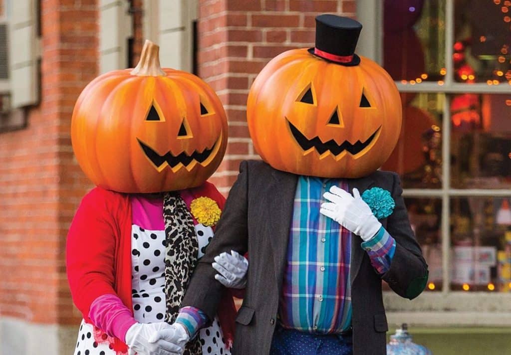 Two people with pumpkin heads, wearing colorful clothing, including a polka-dotted blouse and plaid shirt, stand arm in arm in front of a brick building.