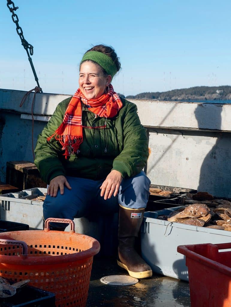 Person sitting on a boat surrounded by baskets and seafood, wearing a green jacket, orange scarf, and boots, smiling in a sunny outdoor setting.