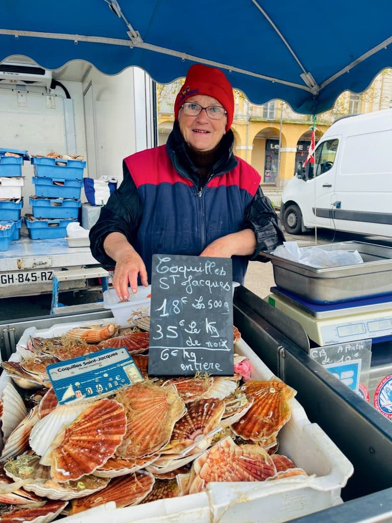 A vendor in a red beanie and glasses stands at a seafood stall displaying scallops with handwritten price signs.