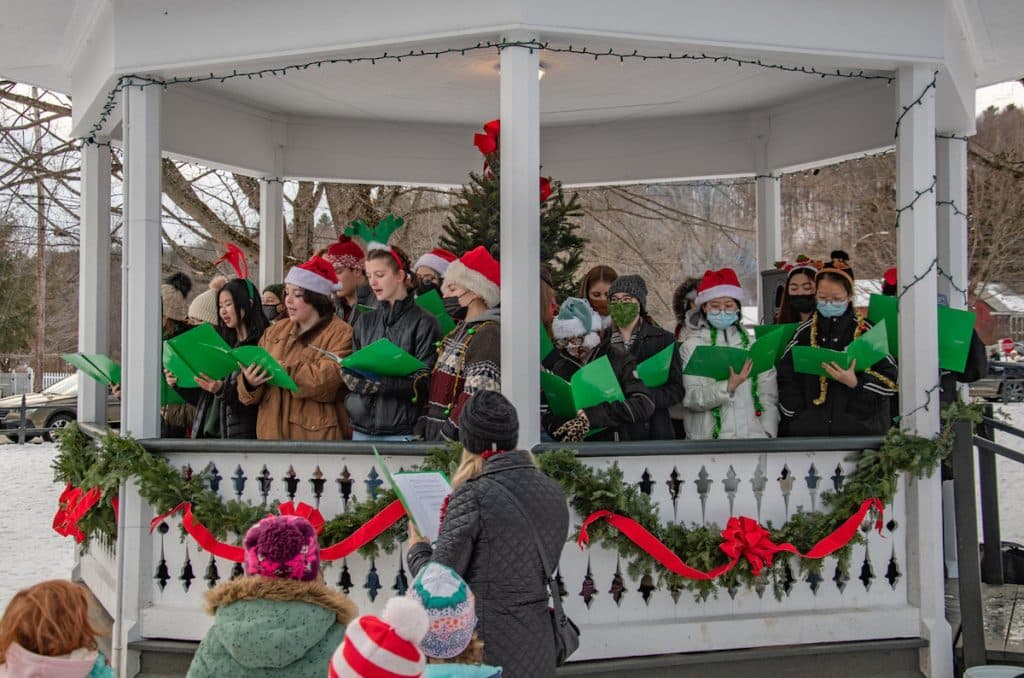 A group of people wearing festive attire sing from green songbooks in a decorated gazebo during a winter event.