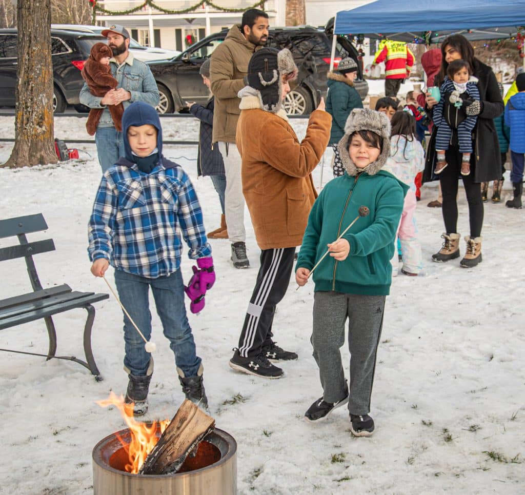 Children roast marshmallows over a firepit in a snowy outdoor event while others gather nearby under a tent.